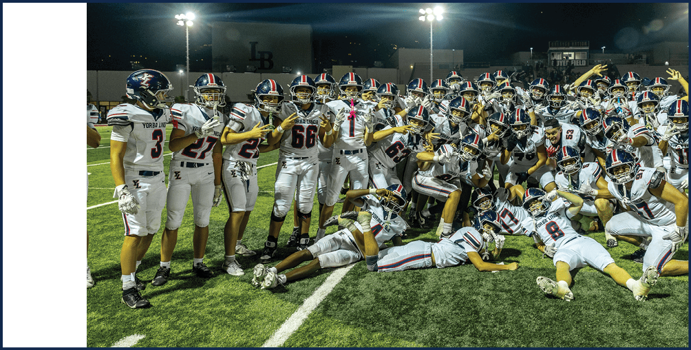 A large group of people wearing matching football uniforms and helmets are gathered closely together on a field, posing energetically with various hand gestures and expressions of excitement. Description generated by AI