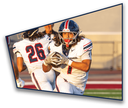 A person wearing a football helmet and uniform is looking forward with a focused expression while another person in a white cap stands blurred in the background. Description generated by AI