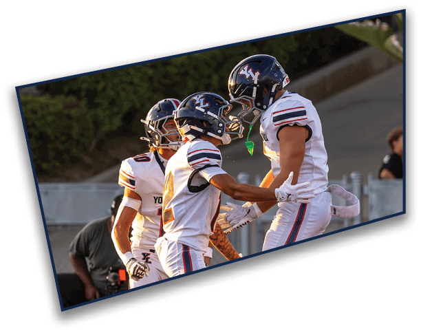 Three people wearing football helmets and white jerseys are closely gathered outdoors with greenery in the background. Description generated by AI