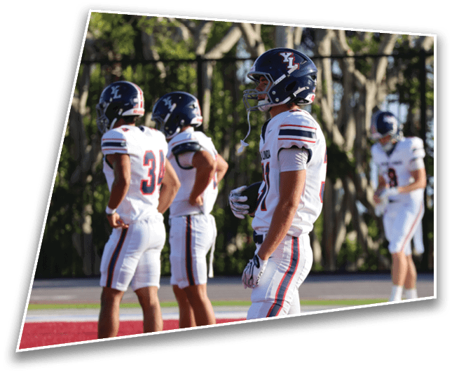 Three people wearing dark blue football helmets with white and red stripes stand outdoors with trees in the background. Description generated by AI