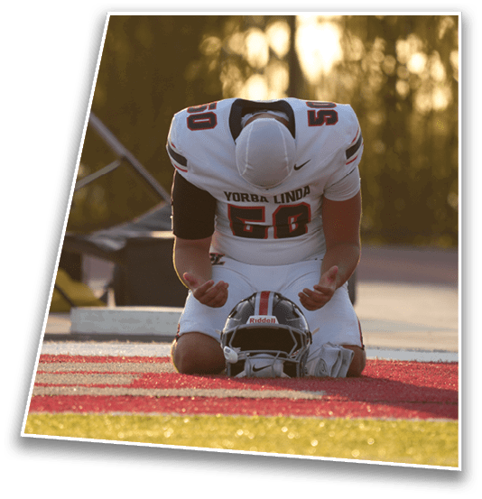 A person in a football uniform kneels on the field with their head bowed and fists clenched near a helmet placed on the ground. Description generated by AI