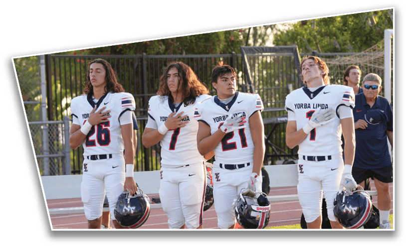 Four people in football uniforms stand on a field with their hands over their hearts holding helmets, accompanied by a person in casual clothing in the background. Description generated by AI