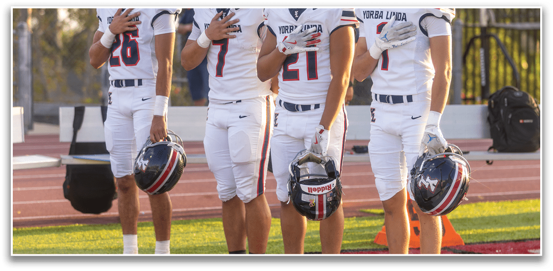 Four people in football uniforms stand on a field with their helmets in one hand and their other hand over their hearts. Description generated by AI
