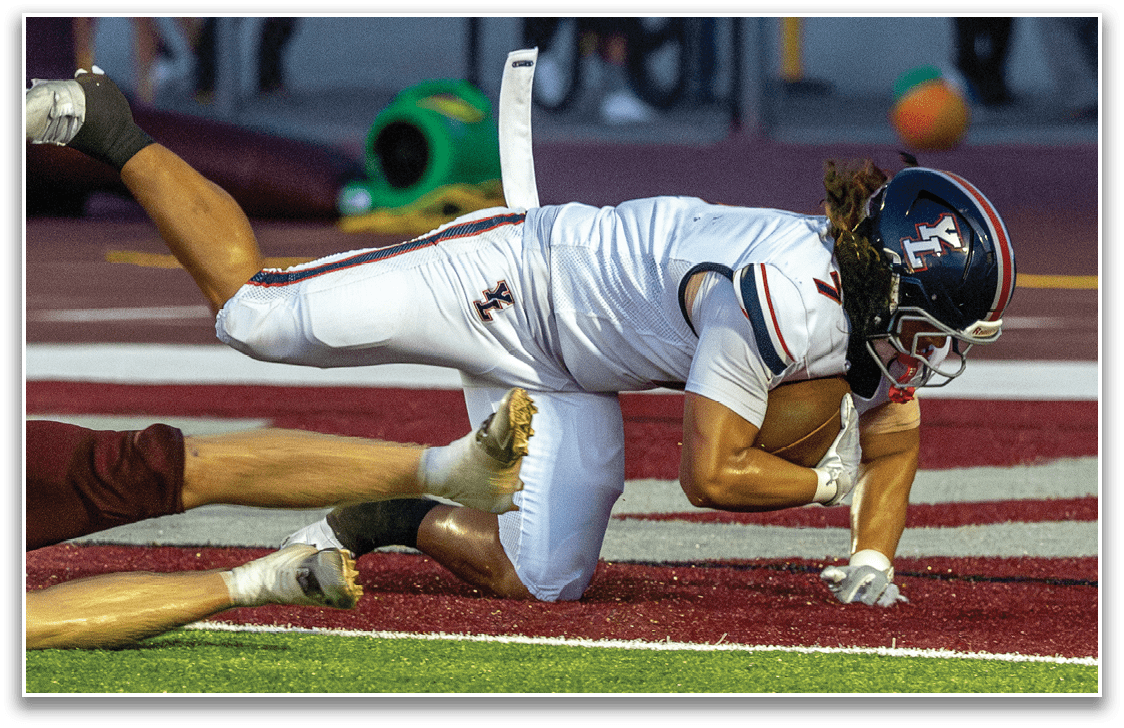 A person in a white football uniform is diving forward while holding a football as another person in a maroon uniform attempts to tackle them on a field near the end zone. Description generated by AI
