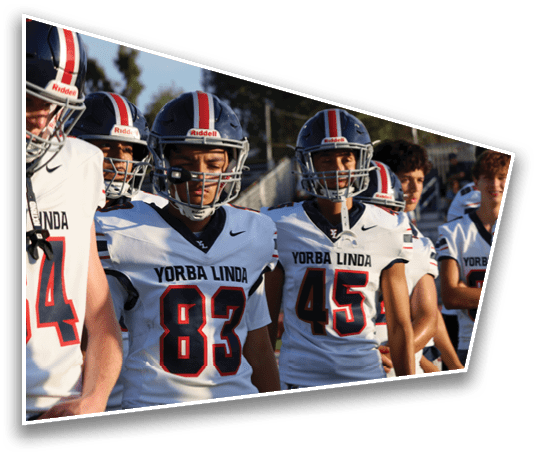 A group of people wearing football helmets and white jerseys with “Yorba Linda" written on them stand together outdoors in sunlight. Description generated by AI