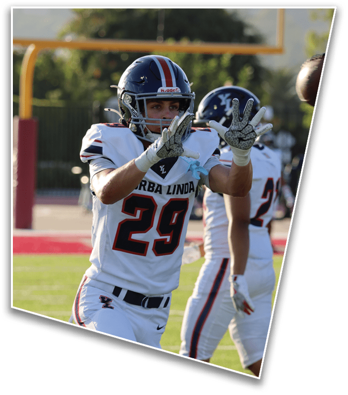 A person wearing a football helmet and gloves is holding up four fingers on one hand with a blurred football field and another player in the background. Description generated by AI