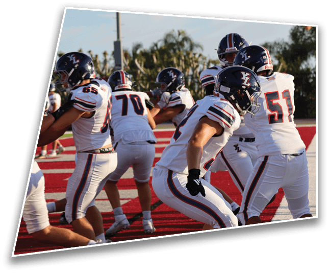 A group of people wearing football uniforms and helmets are gathered on a red field, appearing to prepare for a play or practice. Description generated by AI