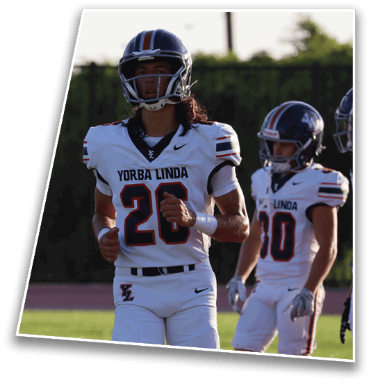 A person wearing a football helmet and a white Yorba Linda jersey with the number 26 stands outdoors with a blurred green background. Description generated by AI
