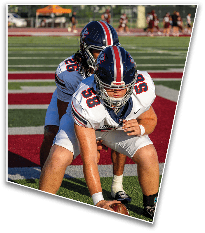 Two people in football uniforms and helmets are positioned on a field with one crouching in front and the other standing closely behind. Description generated by AI