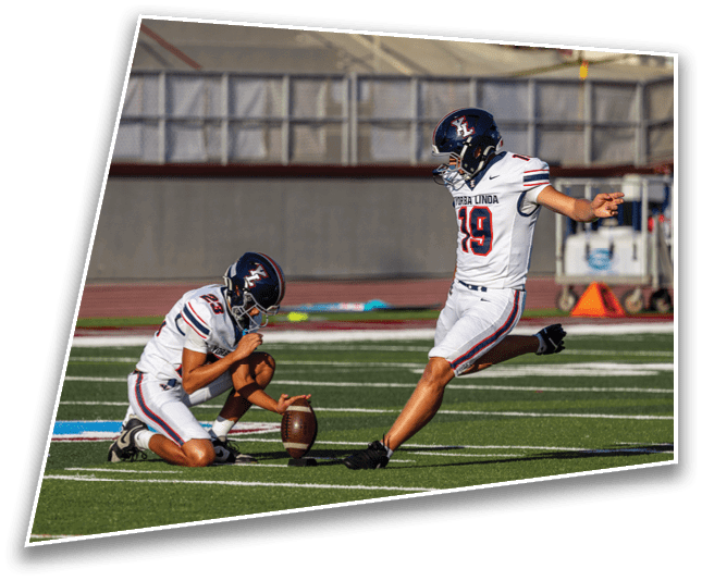 Two people in football uniforms are on a field with one holding a football upright while the other prepares to kick it. Description generated by AI