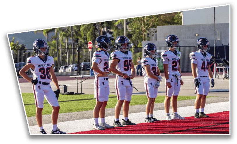 Six people wearing football uniforms and helmets stand in a line on a field near a red carpeted area. Description generated by AI