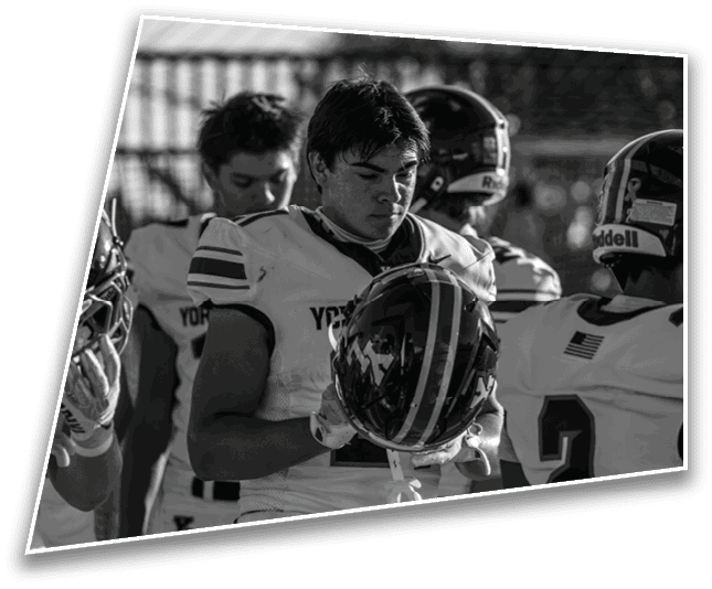 A group of people wearing football uniforms and helmets are gathered closely, with one person holding a helmet and looking down thoughtfully. Description generated by AI