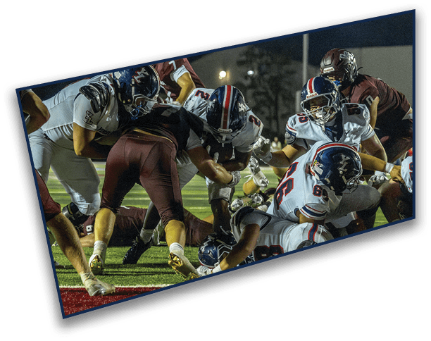Several football players in helmets and uniforms are engaged in a close, intense play on a grassy field under stadium lights. Description generated by AI