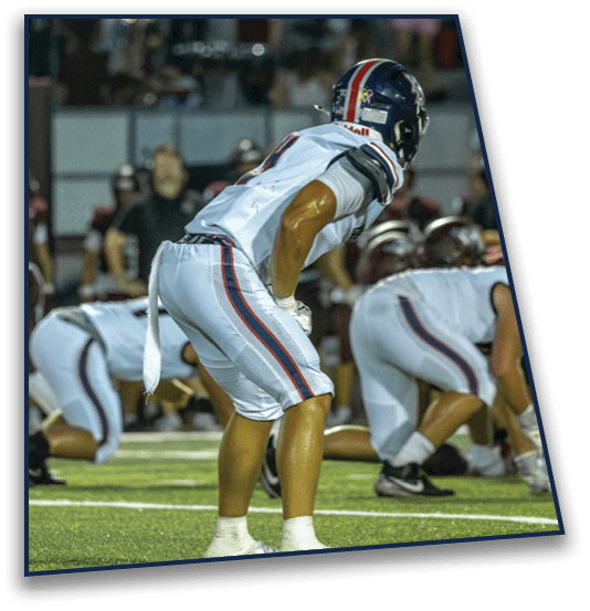 A person wearing a football helmet and white jersey is leaning forward on a field during a game with a blurred crowd in the background. Description generated by AI