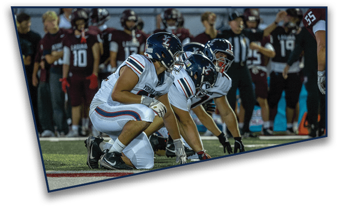 Three football players in white uniforms and helmets are crouched on the field in a ready position while players in dark uniforms stand in the background. Description generated by AI