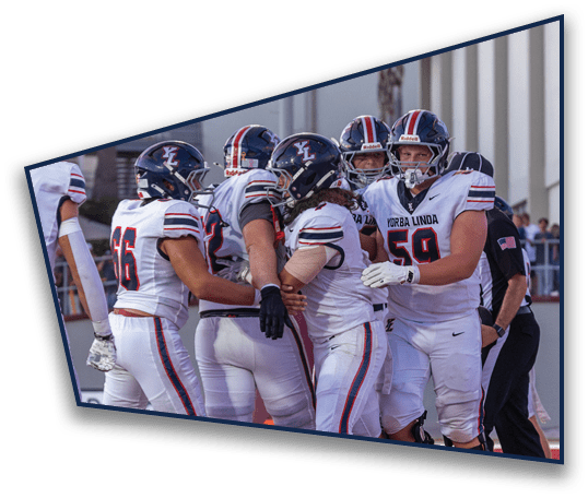 A group of people wearing football helmets and white jerseys with “Yorba Linda" on them are gathered closely on a field near a building with palm trees in the background. Description generated by AI