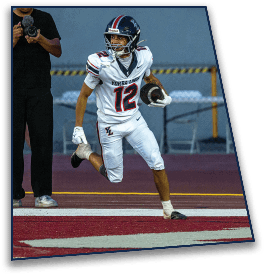 A person in a football uniform with the number 12 is running on a field holding a football while another person stands nearby holding a camera. Description generated by AI