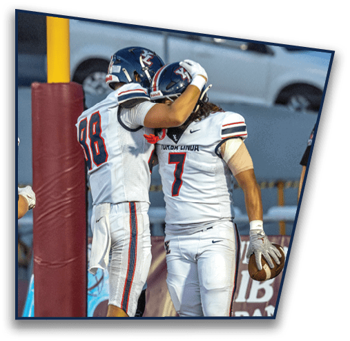 Two people in white football uniforms are celebrating on a field, with one holding a football near a padded goalpost. Description generated by AI
