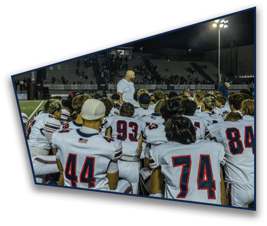 A group of people wearing white sports uniforms with numbers are seated on a field facing a person standing and speaking to them under stadium lights at night. Description generated by AI