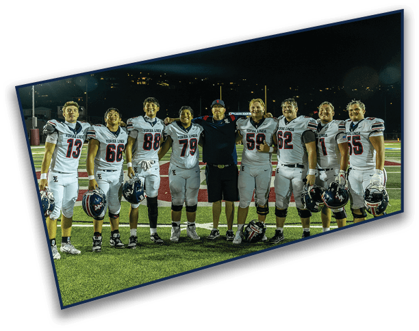 A group of eight people wearing football uniforms and one person in casual clothing stand together on a football field at night, smiling and posing for the photo. Description generated by AI
