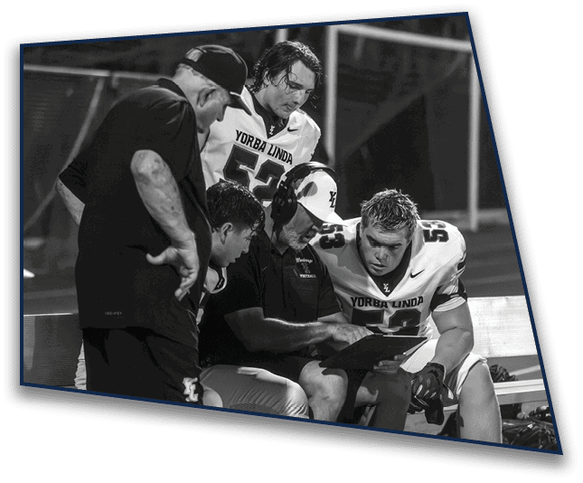 A group of people in football uniforms and a coach are gathered closely, seemingly discussing strategy during a game at night. Description generated by AI