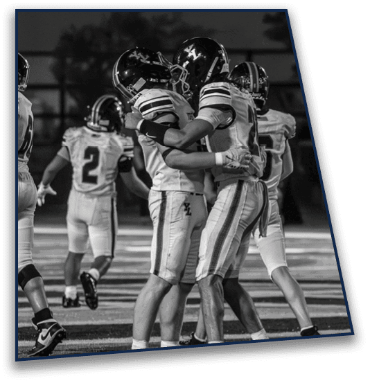 Two people in football uniforms and helmets embrace on a field while other players stand nearby. Description generated by AI