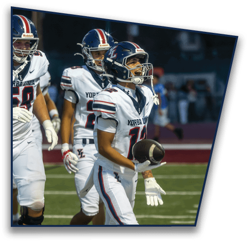Three people in football uniforms with helmets are on a field, with one person holding a football and appearing to be celebrating. Description generated by AI