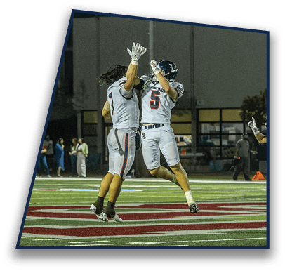 Two people in football uniforms jump to high-five on a field while another person in uniform watches nearby. Description generated by AI