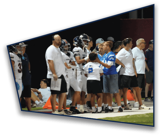 People in sports uniforms and casual clothing stand and interact on the sidelines of a football field during a game. Description generated by AI