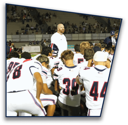 A group of football players in white uniforms are kneeling on a field while two adults stand and address them with a crowd visible in the background. Description generated by AI