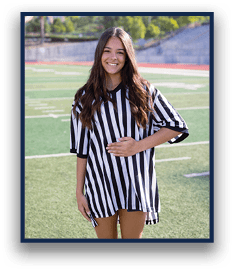 A person with long wavy hair is smiling and standing on a sports field wearing a black and white striped referee shirt. Description generated by AI