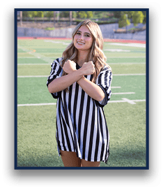 A person with long wavy hair wearing a black and white striped referee shirt stands on a sports field with arms crossed in front of their chest. Description generated by AI