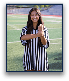 A person with long hair wearing a black and white striped referee shirt is smiling and holding one arm with the other on a sports field. Description generated by AI