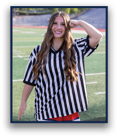 A person with long wavy hair wearing a black and white striped shirt and a colorful skirt is standing on a sports field smiling and posing with one arm bent and fist near their head. Description generated by AI