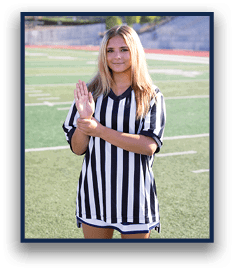 A person wearing a black and white striped referee shirt is standing on a sports field holding one hand up with the other hand gripping the wrist. Description generated by AI