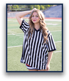 A person with long wavy hair wearing a black and white striped referee shirt stands on a sports field with one hand resting on their head and a slight smile. Description generated by AI