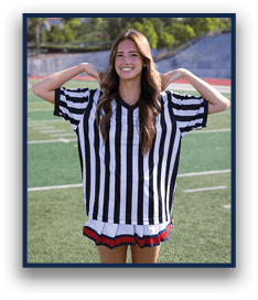 A person with long wavy hair is smiling and posing with hands behind their head while wearing a black and white striped referee shirt and a pleated skirt on a sports field. Description generated by AI