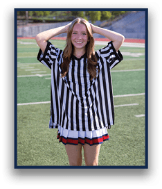 A person with long hair wearing a black and white striped referee shirt and a pleated skirt stands on a sports field with hands resting on their head, smiling. Description generated by AI