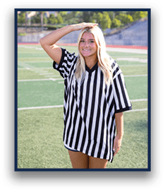 A person with long light hair wearing a black and white striped referee shirt stands on a sports field with one hand on their head and a slight smile. Description generated by AI