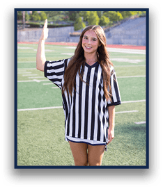 A person with long hair wearing a black and white striped referee shirt is standing on a sports field with one arm raised. Description generated by AI