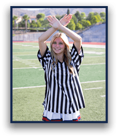 A person with long hair wearing a black and white striped referee shirt is standing on a sports field with arms crossed above their head and smiling. Description generated by AI