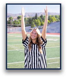 A person wearing a black and white striped referee shirt is standing on a sports field with arms raised and smiling. Description generated by AI