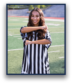 A person with long wavy hair wearing a black and white striped shirt is smiling and crossing their arms in front of their chest while standing on a sports field. Description generated by AI