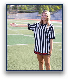 A person wearing a black and white striped referee shirt stands on a sports field with one arm extended to the side and is smiling. Description generated by AI