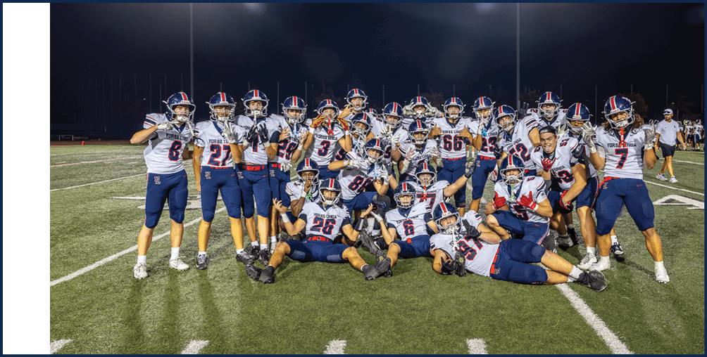 A group of people wearing football uniforms pose together on a lit football field at night, some standing and others sitting or lying on the grass. Description generated by AI