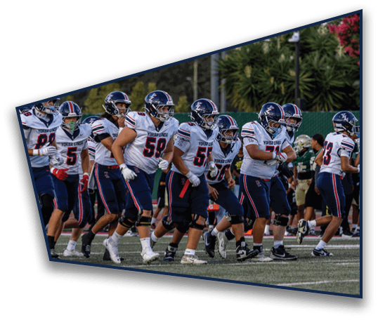 A group of people wearing football uniforms and helmets are running together on a field with trees and a fence in the background. Description generated by AI