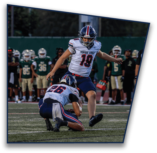 Two people in football uniforms are on a field with one holding the ball on the ground and the other preparing to kick it during a game. Description generated by AI