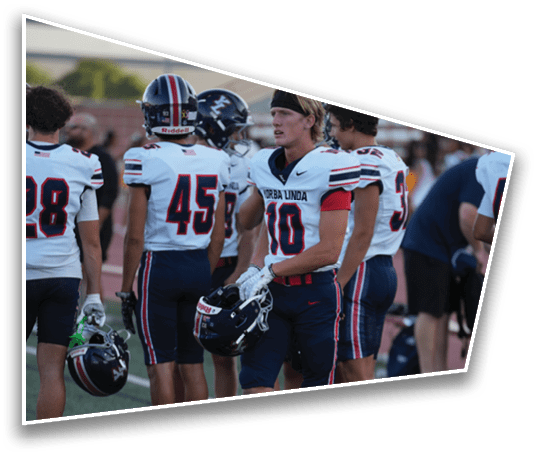 Four people wearing football uniforms with helmets and gloves are standing together on a field with bleachers in the background. Description generated by AI