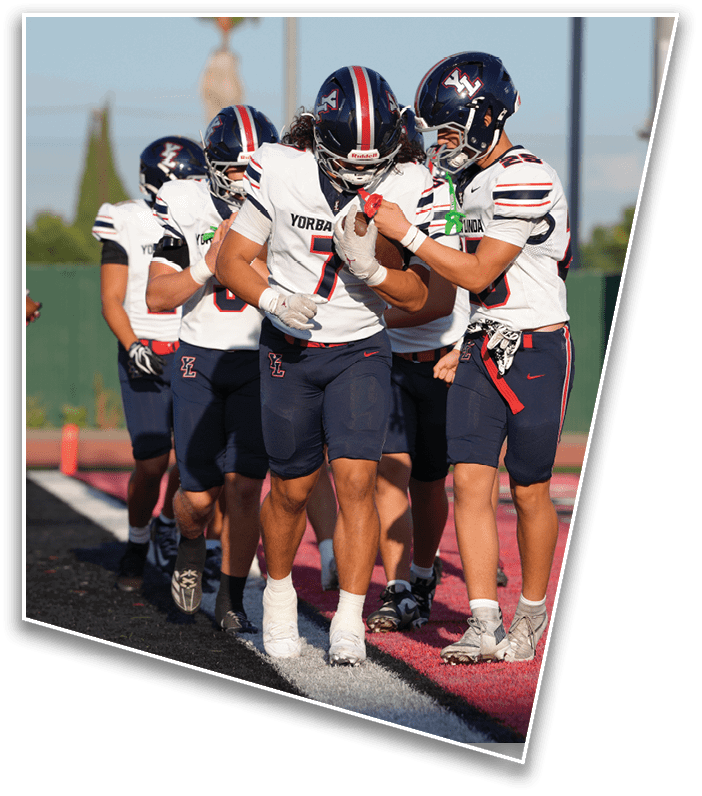 A group of people wearing sports uniforms are standing and walking on a football field near the end zone with red and black turf. Description generated by AI