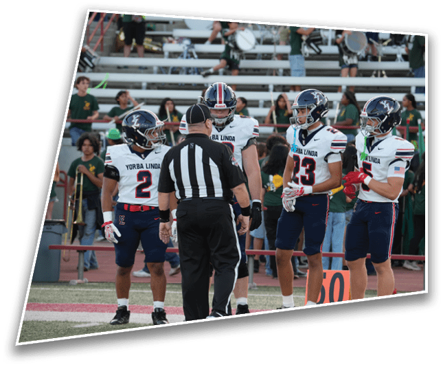 Four people in sports uniforms and a referee stand on a football field near a yard marker. Description generated by AI